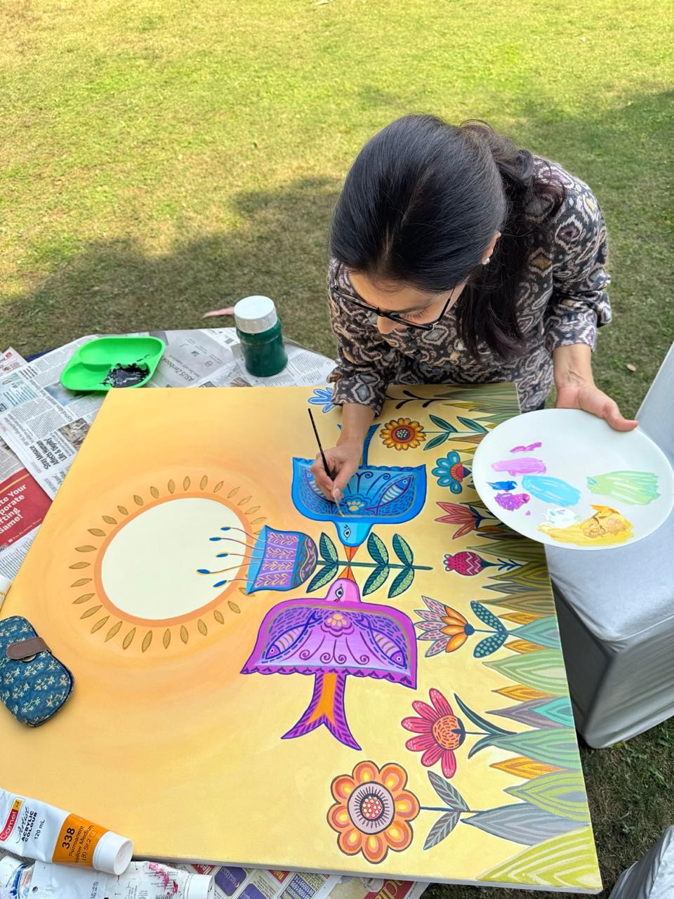 An overhead view of Pavit adding fine details to a blue bird on a golden canvas, holding a palette of paint.